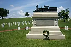 A white marble monument shaped like an altar with steps, with on top a bronze model of a steam engine, on a grassy cemetery with white headstones in the background