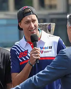 A man in his late twenties wearing a baseball cap the opposite way and a blue and white T-shirt. He is holding a black microphone in his right hand and is speaking to the press