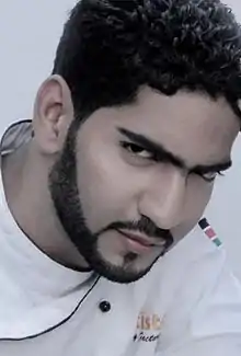 A man with dark hair and beard wearing a white tunic with head tilted and squinting at the camera