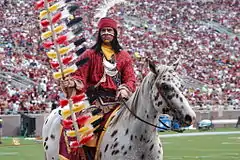 A brown and white spotted horse ridden by a sports mascot in modern-day Native American attire waving a flag stands on a sports field. More people are visible on the field, and a large crowd fills the stadium seating in the background.