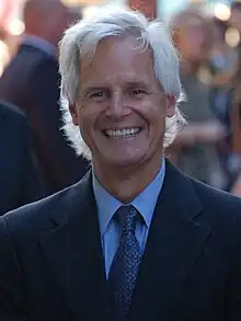 A man with white hair looks into the camera and smiles.