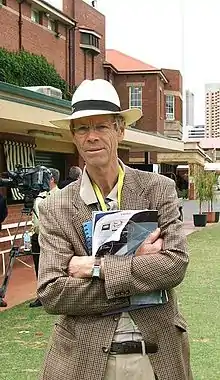  A man in a tweed jacket and a wide-brimmed hat, with his arms crossed holding some papers