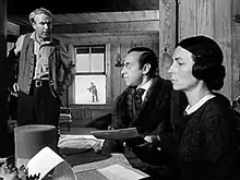 A stern looking man and a woman sit on the right side of a table with documents on the table. A top hat is on the table. An unkempt man stands to the left of the picture. In the background a boy can be seen through a window playing in the snow.