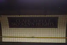 A dark tile mosaic on the curved wall of the station, reading "Clark Street Brooklyn Heights". The rest of the wall is clad with white tile.