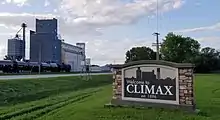 Sign at the side of a state highway reading "Welcome to Climax, established 1896". Large grain elevators are visible in the background.