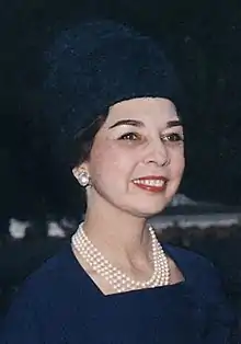 A smiling woman with fair skin and dark hair, wearing a dark blue dress and matching hat, and pearl earrings and necklace