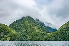 Densely forested mountains rise from the sea, with clouds obscuring their peaks.