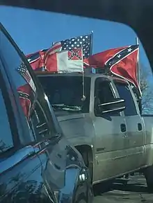 A large pickup truck has three clearly visible flags mounted onto it. The modern US flag, the Confederate Battle flag, and a Confederate National flag.