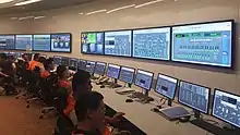 People in hi-vis jackets sitting at a long desk with displays and larger displays on the walls behind