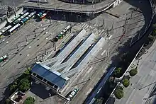 A parking lot filled with buses, seen from above. A station with several blue-roofed platforms can be seen towards the bottom, with lanes for buses and tracks for trains.