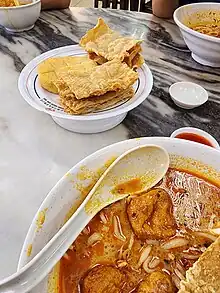 Image 79A bowl of curry mee, with fried beancurd skins and fish cake on the side (from Malaysian cuisine)