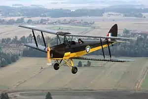  Colour photograph of a black and white biplane flying above a rural area.