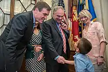 Two older white men in suits bend down as one shakes the hand of a young boy.