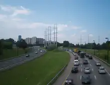 A six lane freeway curving to the right. The opposing directions of travel are separated by a grassy median. Power lines cross the highway, and apartments are visible in the background