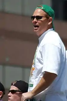 A man, wearing a green hat and white shirt, is standing on top of a parade float in celebration of winning a championship.