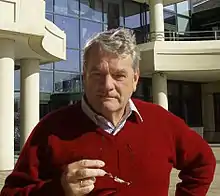 A colour photograph of a grey-haired, thick-set man in his sixties looks directly at the viewer. He wears an open-necked short and red pullover, and holds his spectacles in front of him.