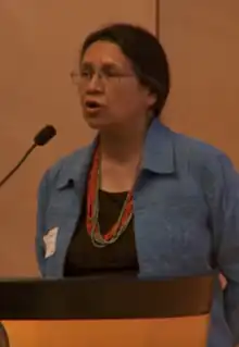 Reese stands behind a podium and is speaking into a microphone. She has dark hair pulled back from her face, light brown skin, glasses, and is wearing a blue jacket over a black shirt, and turquoise and red beaded necklaces.