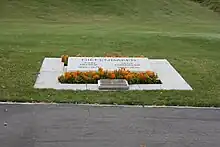 A grave marked by an angled slab of marble engraved with the names of Diefenbaker and his wife and surrounded by plantings of small marigolds, and an additional plaque in bronze. Beyond it a lawn stretches away.