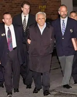 A white-haired man in a black overcoat and dress shirt with his hands behind his back at the center of a small group of men walking toward the camera. The two men on either side are wearing jackets with gold badges clipped to the lapels and ties. They are holding the arms of the man in the center. A fourth man, also in a jacket and tie, is visible in the rear.