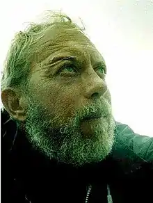 Close-up photo of the rugged face of a man, with grey hear, medium-length grey beard and a long scar across his right eyebrow, facing to right of frame.