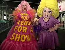 A photograph of two people; on the left is a female-presenting individual with pink hair wearing a pink dress with the text "Are you ready for a show?", and on the right is someone wearing a costume with blonde hair and a purpose dress with the text "Drag is magic".