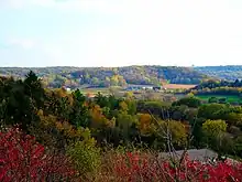 Landscape with low rolling hills, farm buildings in the middle distance surrounded by fields, and bright early autumn foliage