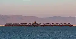 A rust-covered bridge crosses over calm water, with hills and mountains in the background.