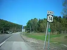 Ground-level view of a road and an associated road sign. More roadsigns and an overhead bridge are visible in the distance.