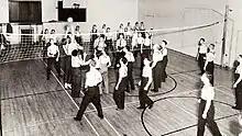 Black and white image of young men dressed in white shirts and dark pants volleying a ball over a net