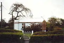 Small light coloured building behind a hedge and gate.