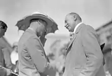 The same mustachioed man, right, wearing a light gray suit and smiling while shaking hands with a man, left, in a light gray suit with a flat-top straw hat and a more bemused expression.