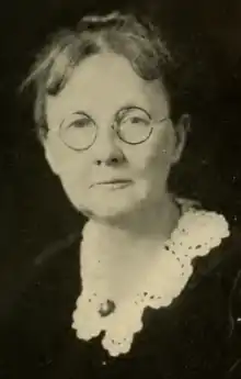 An older white woman, wearing round glasses and a white lace collar