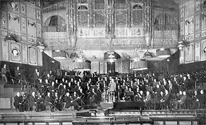 large orchestra and their conductor seen on the platform of Victorian concert hall in long shot