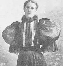 Photograph of a bespectacled woman with her hair pulled away from her face, wearing a frilled high-necked blouse with extremely large puffed sleeves and a striped bodice.