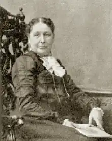 B&W portrait photo of a seated, middle-aged woman with hair in an up-do, wearing a dark dress with a white lacy kerchief, holding a book.