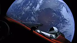 Large circular disc of a fully-illuminated planet Earth floating in the blackness of space. In front of Earth is a red convertible sports-car seen from the side. A humanoid figure wearing a white-and-black spacesuit is seated in the driving seat with the right-arm holding the steering wheel, and the left-arm resting on the top of the car door.