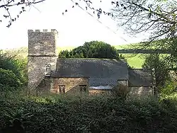 Stone building with tiled roof and square tower, surrounded by vegetation.