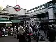 A grey building with a blue sign reading "EMBANKMENT STATION" in white letters and people standing in the foreground all under a white sky