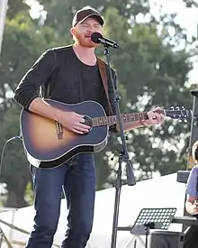 A man with a red beard, wearing a black baseball cap, black t-shirt, and jeans. He is holding a brown guitar and singing into a microphone.