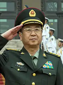 a man standing at attention, wearing a green military uniform, a light green shirt, a dark green tie and a military cap