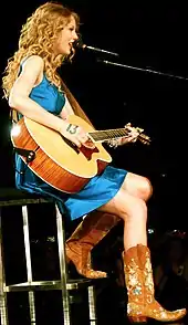 Swift wearing a blue dress and cowboy boots playing a guitar