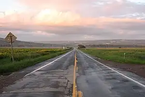 A two-lane paved road, wet in spots, stretching straight ahead with fields on either side towards a ridge in the distance. Above it in the sky is a cloud with orange-tinted sunlight on it; there is also a low mist over the fields in the distance.