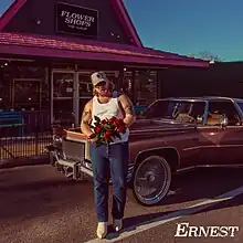 A man in a white tanktop and jeans, holding roses while standing in front of a station wagon. Behind the station wagon is a small storefront with a red roof and a sign reading "Flower Shops (The Album)".