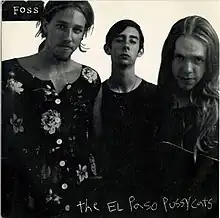 A black-and-white photograph of three young men looking into the camera. Beto O'Rourke, at left, is wearing a floral-pattern dress and sports a shoulder-length hairdo and facial hair.