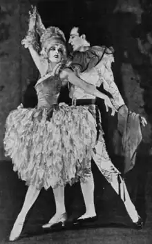 A man and woman in costumes, in a dance pose; she is wearing a skirt that is wide at the hips and ends just below the knees; and he is wearing breeches that tie just below the knee; she is wearing a wig and a corset-style bodice