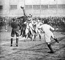 Old cartoon of an association football match with the goalkeeper in the middle who jumps and hits the ball