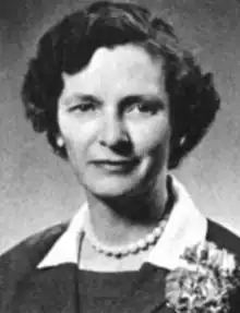 A middle-aged white woman with coiffed hair, wearing white beads and a collared jacket with a corsage