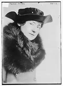 A white woman dressed in a dark fur wrap and a black hat with  an upturned brim.