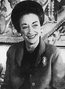 Black and white portrait of a society matron wearing a suit and decorative brooch and sitting in front of an ornate wall decoration