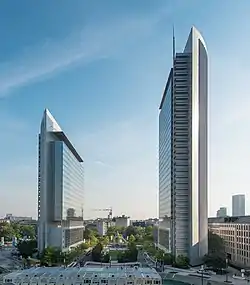 Two skyscrapers stand at either side of the frame. They appear to be made of metal and glass. Pollux, the building on the right, is visibly taller than Kastor on the left. Between the buildings is a grassy median with a metal art piece and trees.